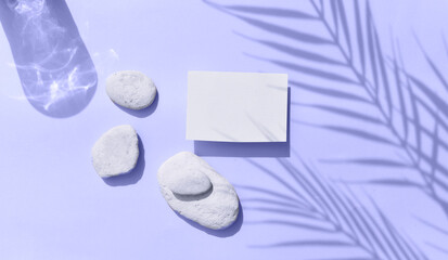 Summer tropical still life mockup composition. Blank invitation card with sea pebbles on lilac background. Harsh palm shadows, reflection.  Mockup empty greeting  blank for wedding or business.
