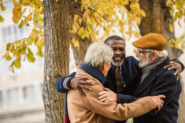 Smiling african american man hugging interracial friends in autumn park.