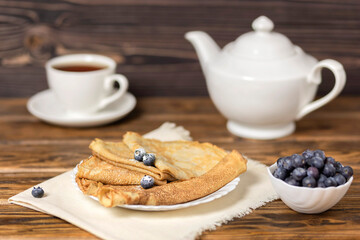 Fried pancakes with blueberries on a wooden table with a white teapot and a mug of tea in a rustic style. Maslenitsa, a classic Russian and Ukrainian dish. Selective focus.