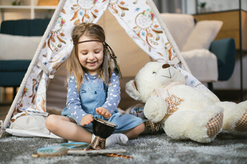 Happy female child with headband in boho style playing on little drum while sitting in wigwam at home. Concept of carefree, childhood and enjoyment. © sofiko14