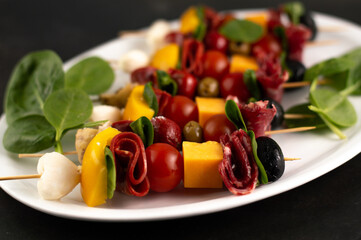 Blurred image of appetizers on skewers with mozzarella, tomatoes, peppers, salami, spinach on a white plate on a dark background.