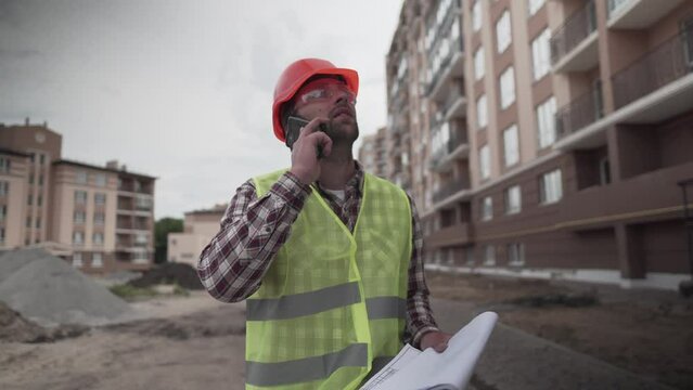 Working process. Contractor in working uniform control according to plan. Builder phone call and walking along construction site of new residential area. Foremant supervising construction.