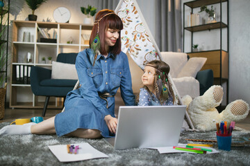 Pleasant mother and her daughter in headbands in boho style sitting together near toy wigwam and using wireless laptop. Concept of family, technology and time spending. © sofiko14