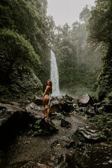 A young woman traveller is exploring a waterfall in tropical settings of Bali islands, Indonesia. Tropical forest, lush greenery, rive