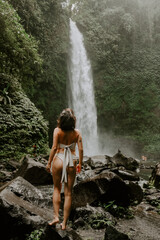 Fototapeta na wymiar A young woman traveller is exploring a waterfall in tropical settings of Bali islands, Indonesia. Tropical forest, lush greenery, river