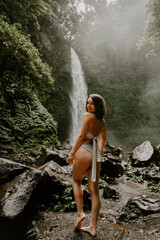 Fototapeta na wymiar A young woman traveller is exploring a waterfall in tropical settings of Bali islands, Indonesia. Tropical forest, lush greenery, rive