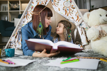 Caring mother and her daughter looking on each other while lying inside toy wigwam with book in hands. Woman and kid wearing indian headbands while playing at home. © sofiko14