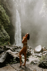 Fototapeta premium A young woman traveller is exploring a waterfall in tropical settings of Bali islands, Indonesia. Tropical forest, lush greenery, river