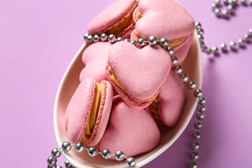 Bowl with tasty heart-shaped macaroons and beads on purple background, closeup