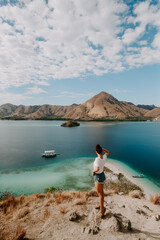 A young female traveler is exploring tropical island in Komodo, Indonesia. She is wearing a casual outfit and overlooks lagoon bag, boat and hills in the distance