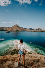 A young female traveler is exploring tropical island in Komodo, Indonesia. She is wearing a casual outfit and overlooks lagoon bag, boat and hills in the distance