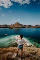 Fototapeten A young female traveler is exploring tropical island in Komodo, Indonesia. She is wearing a casual outfit and overlooks lagoon bag, boat and hills in the distance © Michal