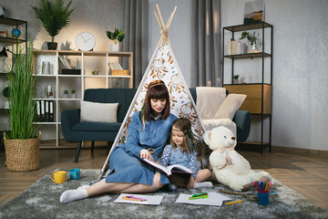 Caring caucasian mother reading fairytale to her pretty daughter while sitting on floor at home. Woman and child wearing headbands in boho style with toy wigwam on background. © sofiko14