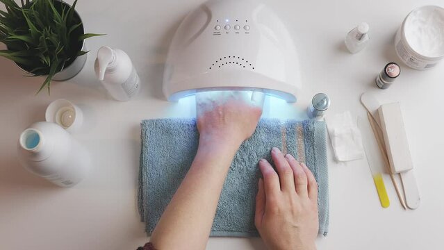 Woman applying gel hybrid polish using UV lamp. Beauty wellness spa treatment concept. Cosmetic products, UV lamp, green leaves on white table. Spa, manicure, skin care concept. Flat lay overhead
