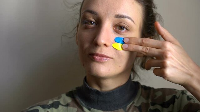 Young girl is paining blue and yellow ukrainian flag on her cheek using her fingers wearing military uniform, mandatory conscription in Ukraine, equality concepts