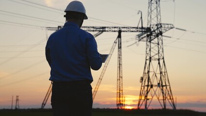 Silhouette Power engineer in white helmet checks power line using computer on tablet, remote control of power system. High voltage electrical lines at sunset. Distribution and supply of electricity
