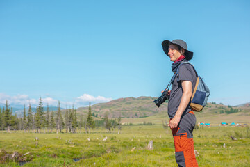 One traveling man with hat and backpack walks in sunlight on vivid bokeh background of coniferous trees and mountain under blue sky. Colorful portrait of tourist with camera in bright sun in mountains