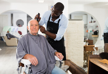 Aged male client getting trendy haircut at barber shop from African-American hairstylist using machine