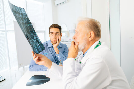 Serious mature adult male physician consult frustrated young man patient giving bad news explaining results of MRI image. Unhappy scared young man listening to bad news sitting in doctor office.