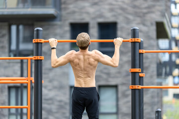 Shirtless muscular man with fit torso having outdoor fitness workout on pull up bar in the street open gym