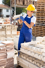 Concentrated young woman employee working in a building materials store carefully examines a brick in a warehouse