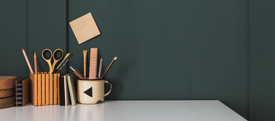 Creative desk with office supplies, on a dark green wall background.	
