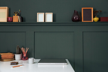 Creative desk with notebook computer, desk objects, office supplies, books, picture frames and cactus on a dark green background.	
