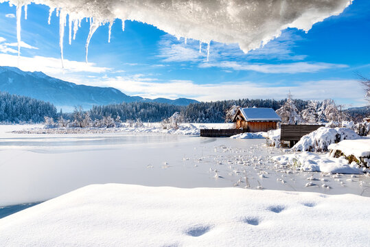 A panoramic view on the Faaker lake in Austrian Alps. The lake is surrounded by high mountains. There is a snow covered pier going into the frozen lake.