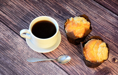 Two fresh cupcakes with sprinkles and a cup of black coffee on a saucer with a spoon on a wooden table.
