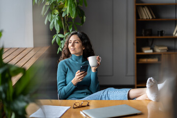 Freelancer writer or social media manager work from home sit cozy at desk with closed laptop and feet on table drink morning coffee holding smartphone in hand. Businesswoman work remotely in mobile