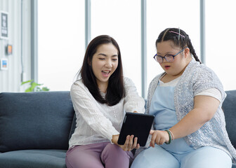 Asian happy lovely young little daughter smiling with mother sitting ,on cozy sofa look at tablet in living room