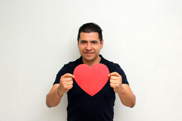 Latin adult man with a big red heart shows his enthusiasm for the arrival of February and celebrate the Day of Love and Friendship with his partner, friends and family sharing affection
