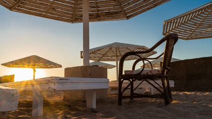 Early morning on the beach of Egypt. Lattice sun umbrellas against the blue sky. Empty sunbeds and a chair stand on the sand. The rays of the rising sun are shining.