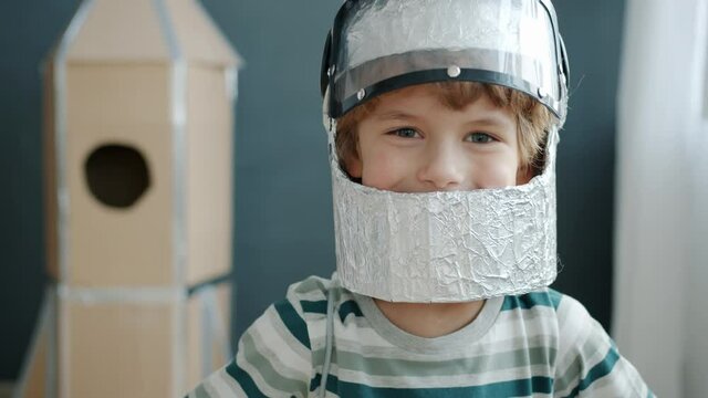 Portrait of adorable blond kid wearing astronaut helmet smiling standing at home against spaceship background. People and positive emotion concept.
