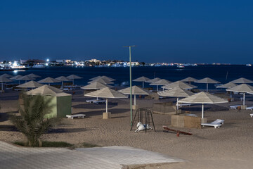 Fototapeta na wymiar Overnight on the Red Sea beach. Sun umbrellas and empty sunbeds stand in rows on the sand. City lights shine in the distance. Deep blue sky. Egypt
