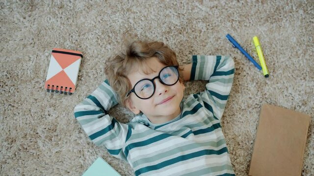 Top view of smart child wearing glasses smiling thinking lying on floor at home with stationery visible around. Genius kids and apartment concept.