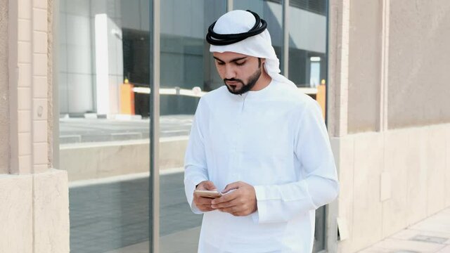 Walking Arab man in Kandura dish dash ideal for Middle East concept. Emirati wearing traditional UAE clothing using mobile phone