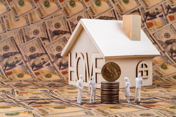 Plastic figurines of symbolic people stand next to a 1 American dollar coin in front of a small house against the background of 100 American dollar bills