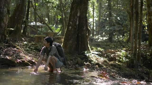 Asian female tourist with backpack washing hands in water stream that flowing through the lush foliage plants in tropical forest. Solo camping. Summer hiking trip in the jungle.