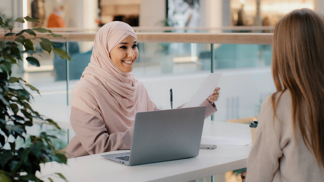 Young arab woman consultant sales agent bank worker sitting at office desk advises unrecognizable girl client offers to sign contract sale agreement helps to arrange loan proposes legal consultation