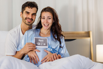 Cheerful husband embracing happy wife on bed at night and showing pregnancy ultrasound image film of unborn baby