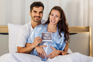 Cheerful husband embracing happy wife on bed at night and showing pregnancy ultrasound image film of unborn baby