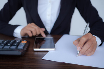 Staff woman holding pen for writing on blank paper and touch screen on mobile or smartphone with calculator on wooden table at office. technology for business or finance concept.