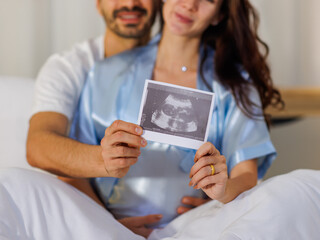 Cheerful husband embracing happy wife on bed at night and showing pregnancy ultrasound image film...