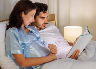 Happy pregnant wife holding belly and relax on bed beside beloved husband and enjoy reading entertainment media on a book together with smile and laugh. Parents expecting joyful life for child birth.