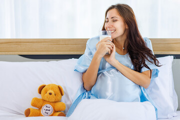 Cheeful pregnant woman smile as enjoy drinking glass of milk beverage in morning while resting on bed with lovely bear doll