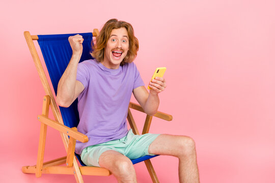 Portrait of handsome trendy cheery lucky guy sitting in chair using device having fun post like isolated over pink pastel color background