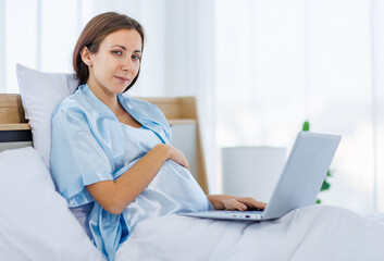 Beautiful pregnant woman  pajamas resting on bed with smile and happy to watch laptop for prenatal care of maternity. Healthy mom relax and pleased with belly of unborn baby for expecting child birth.
