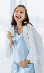 Pregnant woman on night gown happy to drink glass of milk beverage while relaxing near window curtains