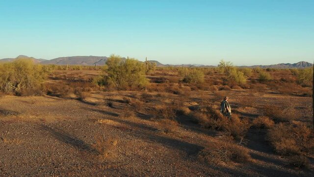 A southwestern desperado man wearing a poncho walks through sagebrush and past towering Saguaro Cacti in the Sonoran Desert at sunset.